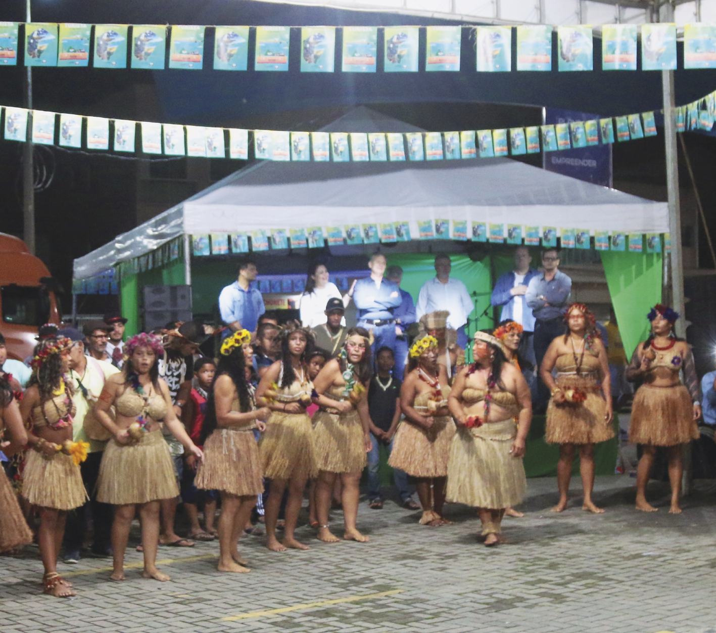 Fotografia. Mulheres indígenas usando saias feitas de palha, coroa de flores amarelas na cabeça e colares seguram chocalhos durante uma apresentação. Ao fundo, uma tenda com pessoas não-indígenas.