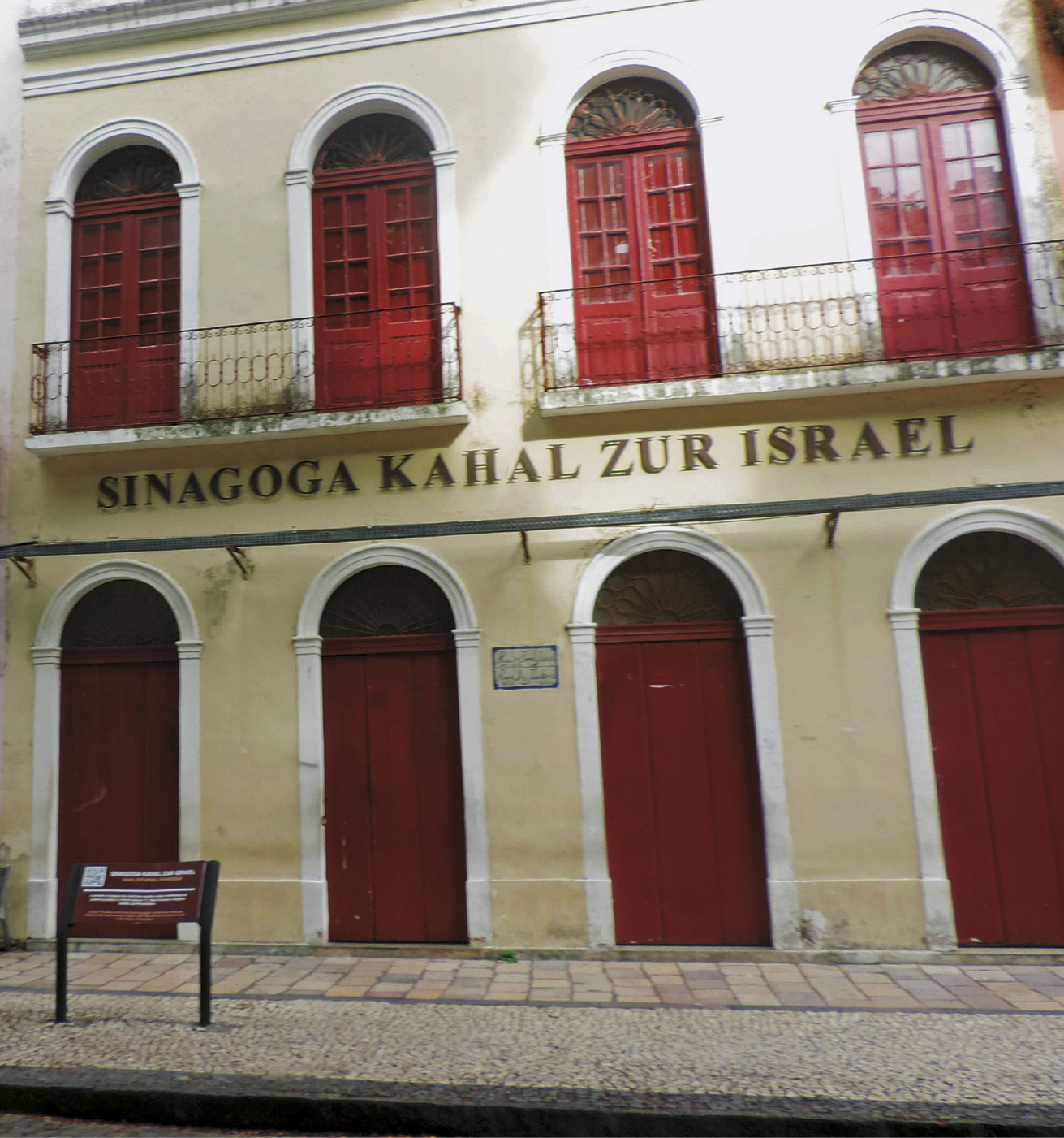 Fotografia. Fachada de edifício amarelo-claro de dois pavimentos com quatro grandes portas vermelhas de madeira em formato de arco no térreo e quatro no andar superior que dão para sacadas. Na frente, a inscrição: Sinagoga Kahal Zur Israel.