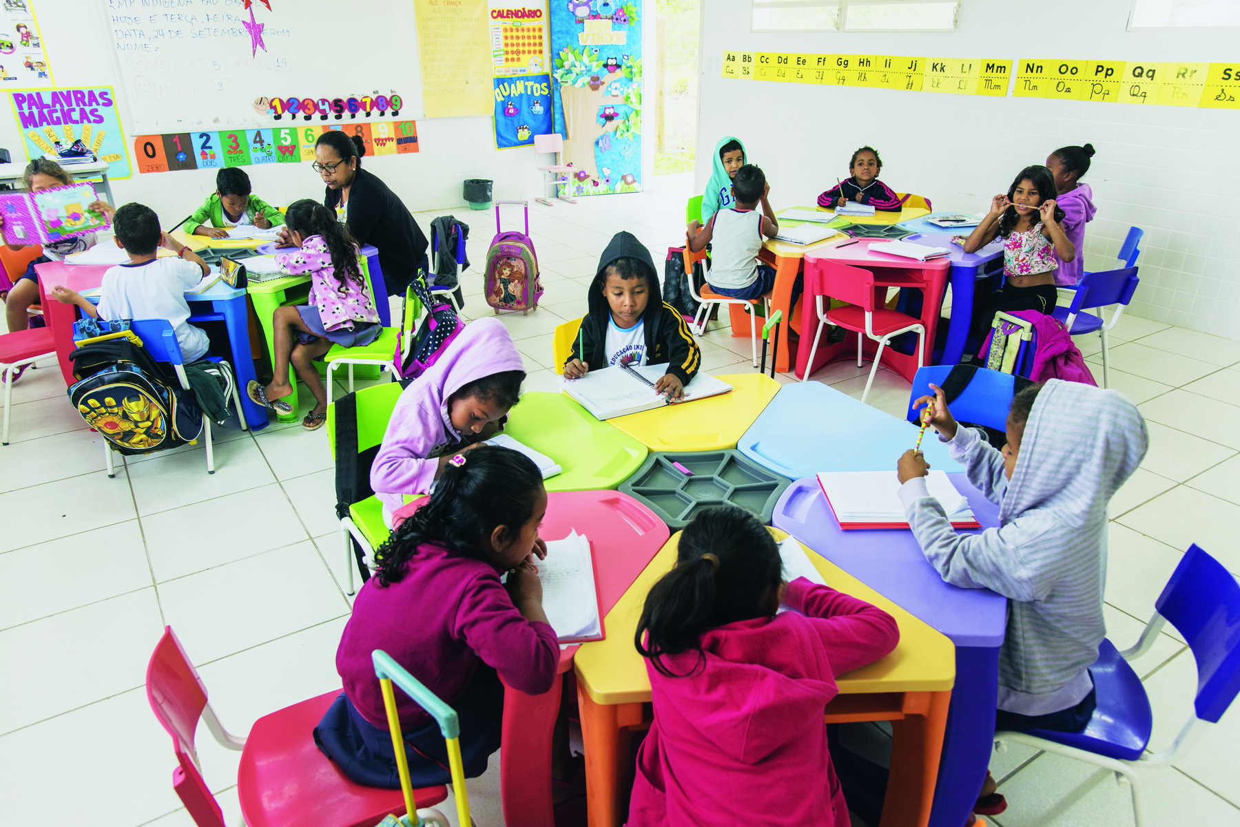 Fotografia. Sala de aula com três mesas coloridas, de forma hexagonal, e crianças sentadas em volta delas. As crianças escrevem em cadernos e, próximo a uma das mesas, à esquerda, uma professora sentada ajuda um grupo de alunos.
