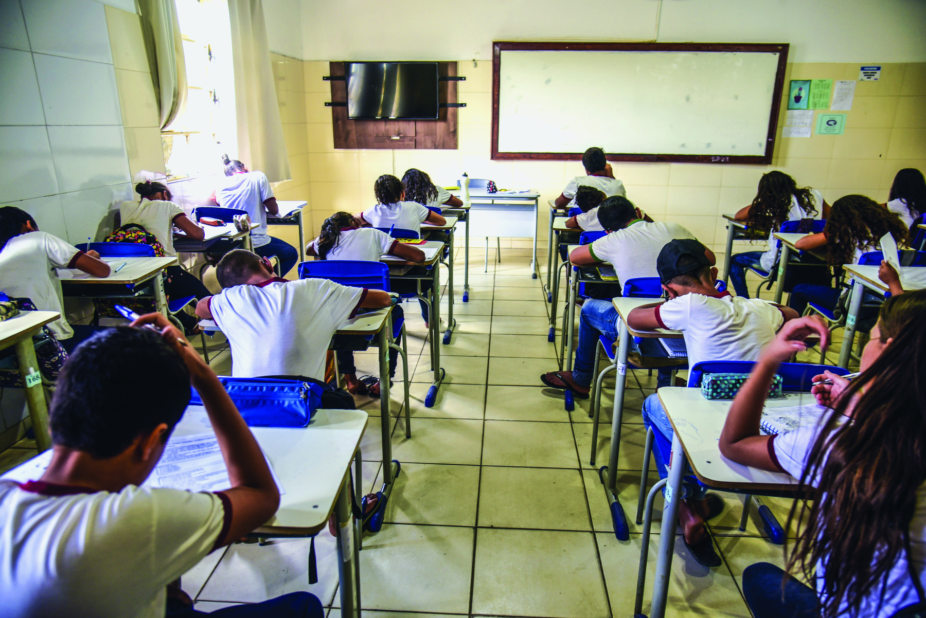 Fotografia. Crianças vistas de costas, sentadas em carteiras enfileiradas, em uma sala de aula. Todas escrevem em folhas de papel.