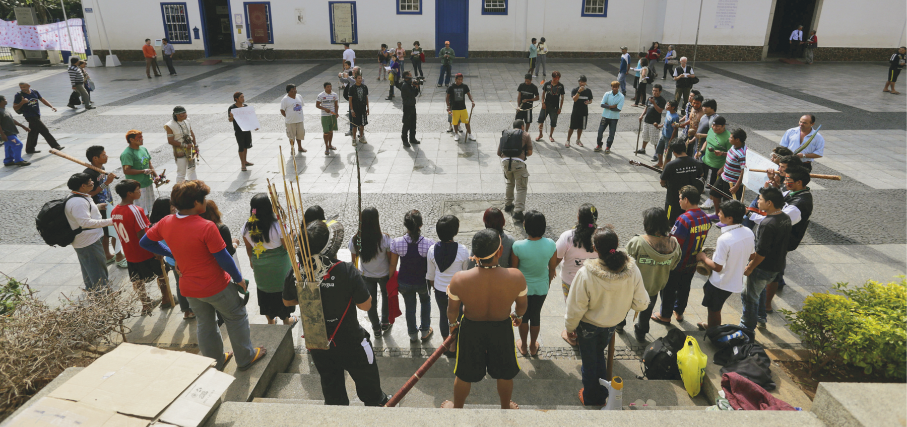 Fotografia. Em um pátio, pessoas indígenas estão reunidas, dispostas em círculo. Algumas portam pedaços de pau na mão, outras, cartazes. Um homem indígena segura um arco e um feixe de flechas. Pessoas ao redor observam o ato.