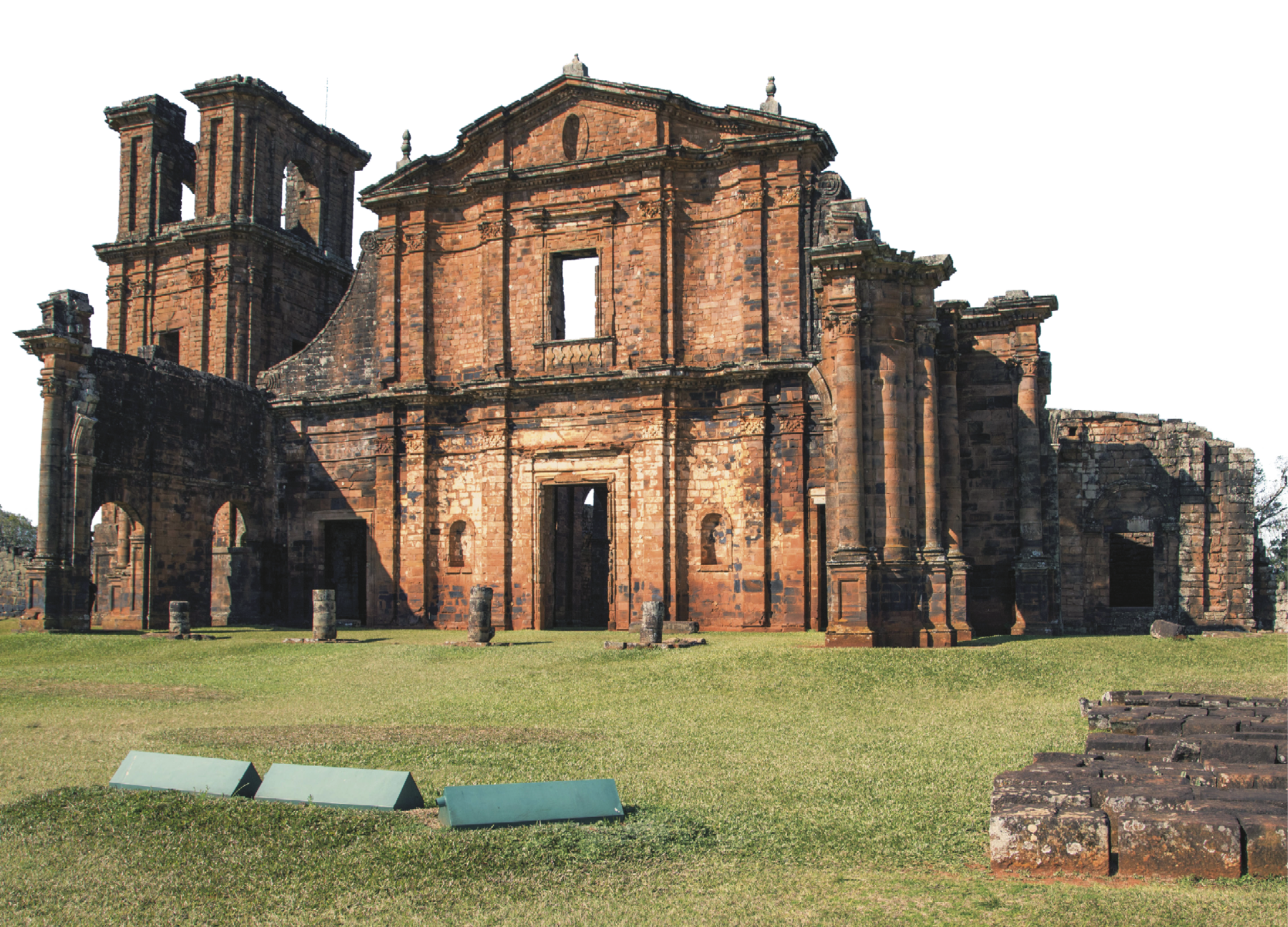 Fotografia. Construção de pedras de cor marrom, formada por colunas nas laterais, à direita, e uma torre quadrada, à esquerda, em meio a um campo plano coberto por grama verde.