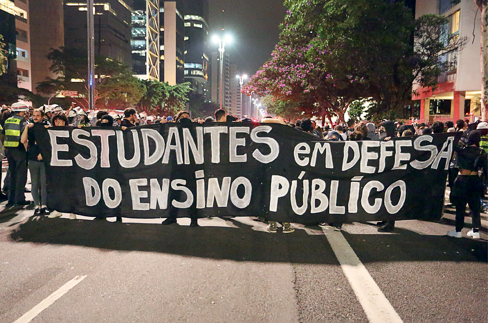 Fotografia. Grupo de pessoas aglomeradas se manifestando em uma larga avenida. À frente uma longa faixa preta com os dizeres: ESTUDANTES EM DEFESA DO ENSINO PÚBLICO.
