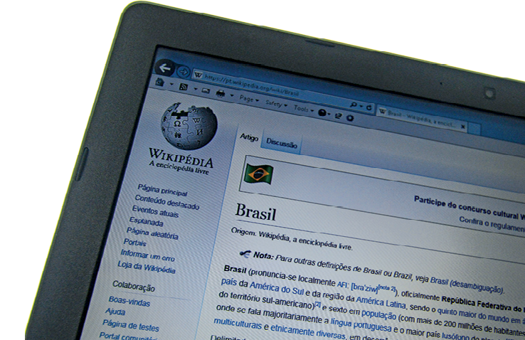 Fotografia. Parte de uma tela de computador que mostra o acesso ao site da Wikipédia. No canto superior esquerdo, o logotipo do site e um menu. No centro da página a bandeira do Brasil, o verbete Brasil e um trecho do texto sobre ele.