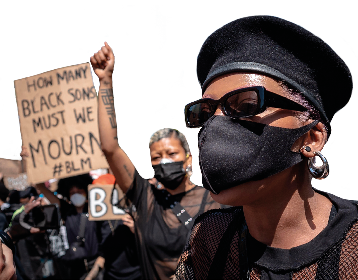 Fotografia. Destaque para duas mulheres negras usando roupas e máscaras de proteção facial, com boca e nariz cobertos. Uma delas usa uma boina. Ao fundo, pessoas carregam cartazes com frases escritas em inglês.