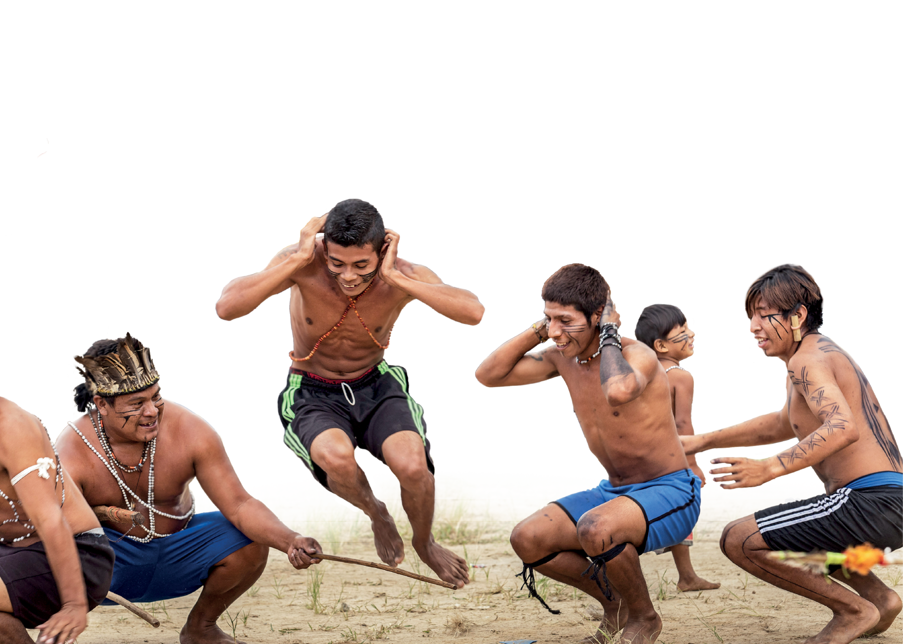Fotografia. Cinco rapazes indígenas de bermuda e com pinturas faciais e colares, estão agachados e com as mãos sobre a nuca. Um deles salta, enquanto um outro, à direita, movimenta uma haste, próxima ao chão. Eles estão sorrindo.