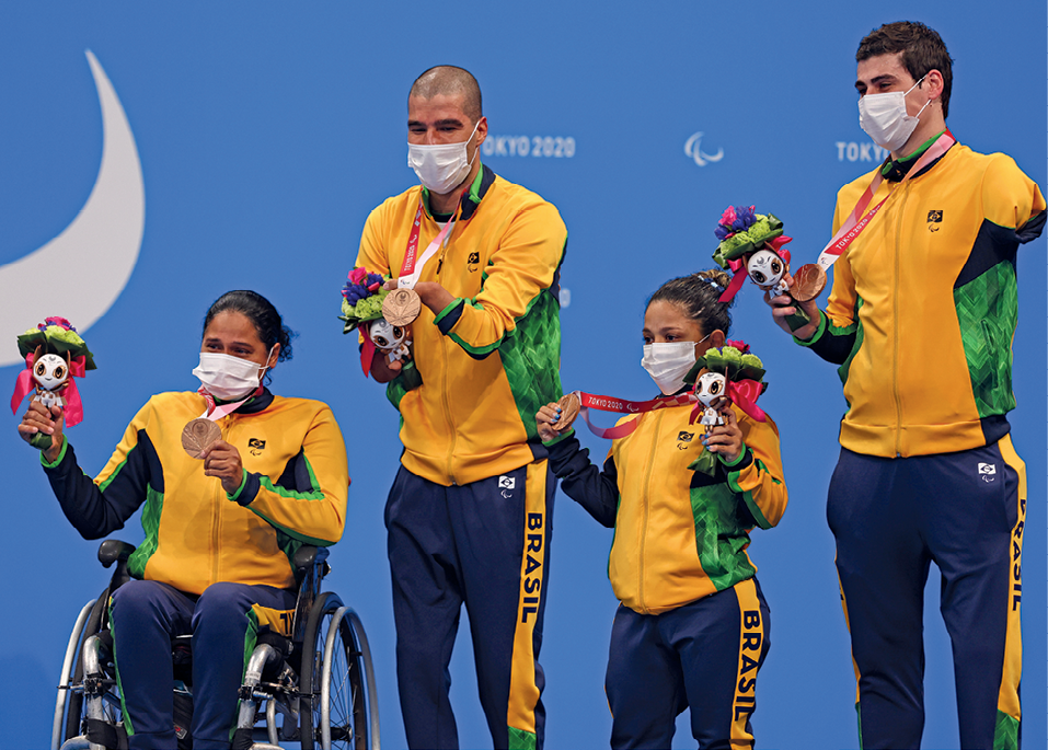 Fotografia. Duas mulheres e dois homens, todos atletas paralímpicos. Eles usam máscara de proteção no nariz e na boca, vestem calças em azul escuro e blusa verde e amarela - o uniforme do Brasil - e seguram um buquê e uma medalha.