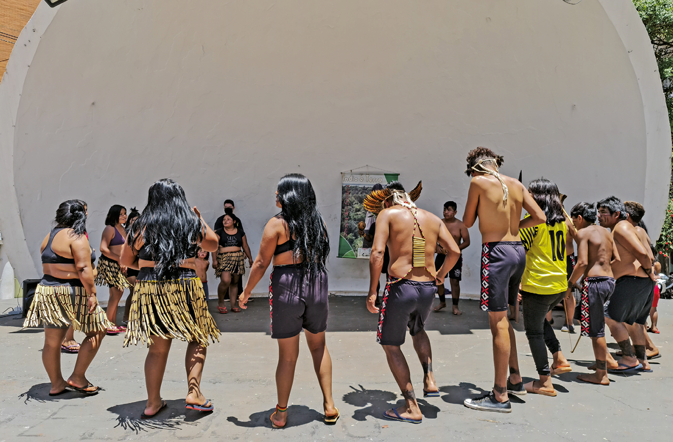 Fotografia. Em um local ao ar livre, com chão asfaltado, um grupo de  indígenas executa uma dança circular na frente de uma construção  de formato semicircular. Os homens usam bermuda e cocar. As mulheres usam bermuda, saia de palha, top e possuem cabelos pretos e compridos. Muitos estão de chinelo. Eles são vistos de costas.