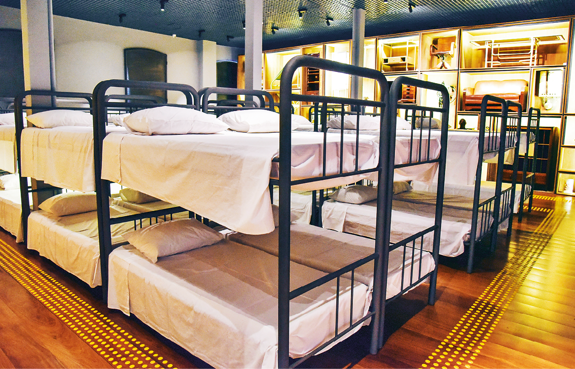 Fotografia. Vista para um amplo salão com piso de madeira e muitas camas beliche de ferro preto agrupadas. As camas estão forradas com lençóis brancos e possuem travesseiros brancos.