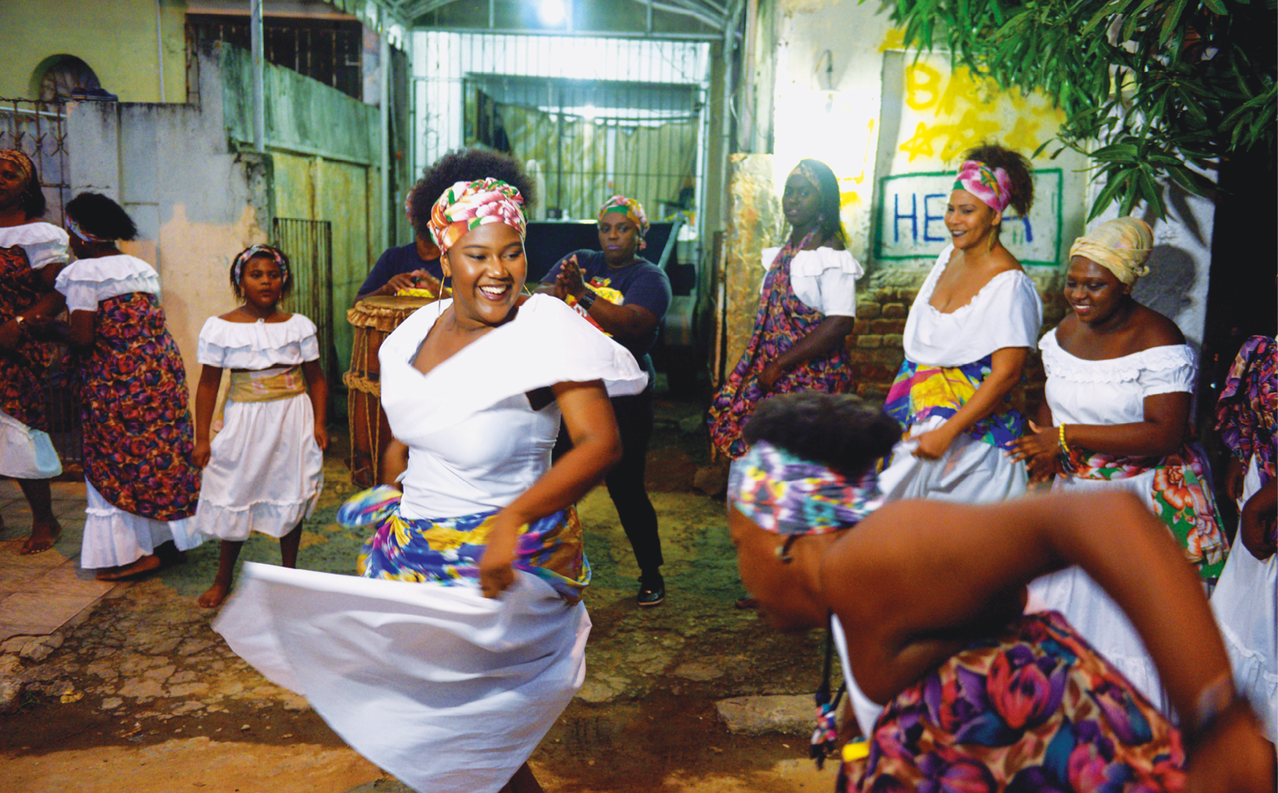 Fotografia. Vista para a calçada de uma rua em frente a uma casa com portão gradeado. Uma dezena de mulheres negras sorridentes dançam em roda. Elas vestem roupas brancas com lenços coloridos na cabeça e na cintura.