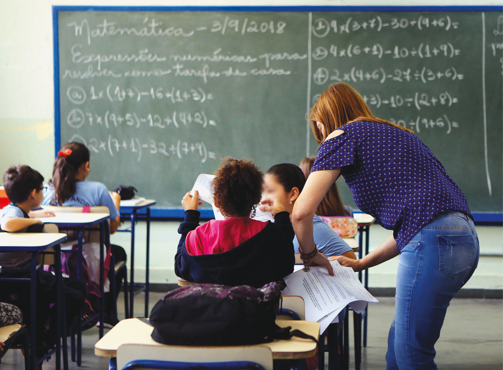 Fotografia. Crianças vistas de costas sentadas em carteiras enfileiradas em uma sala de aula. A professora está em pé, à direita, ao lado de uma das crianças, distribuindo folhas de papel. Ao fundo, lousa com expressões matemáticas.