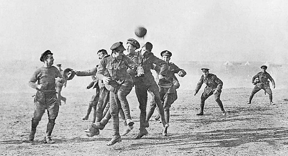 Fotografia em preto e branco. Soldados sorrindo e jogando bola em uma área aberta. No centro dois deles disputam a bola, no alto.