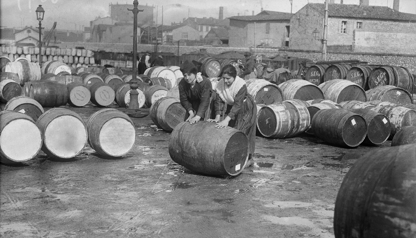 Fotografia em preto e branco. Duas mulheres empurrando um barril deitado. Ao redor delas outros barris no chão.