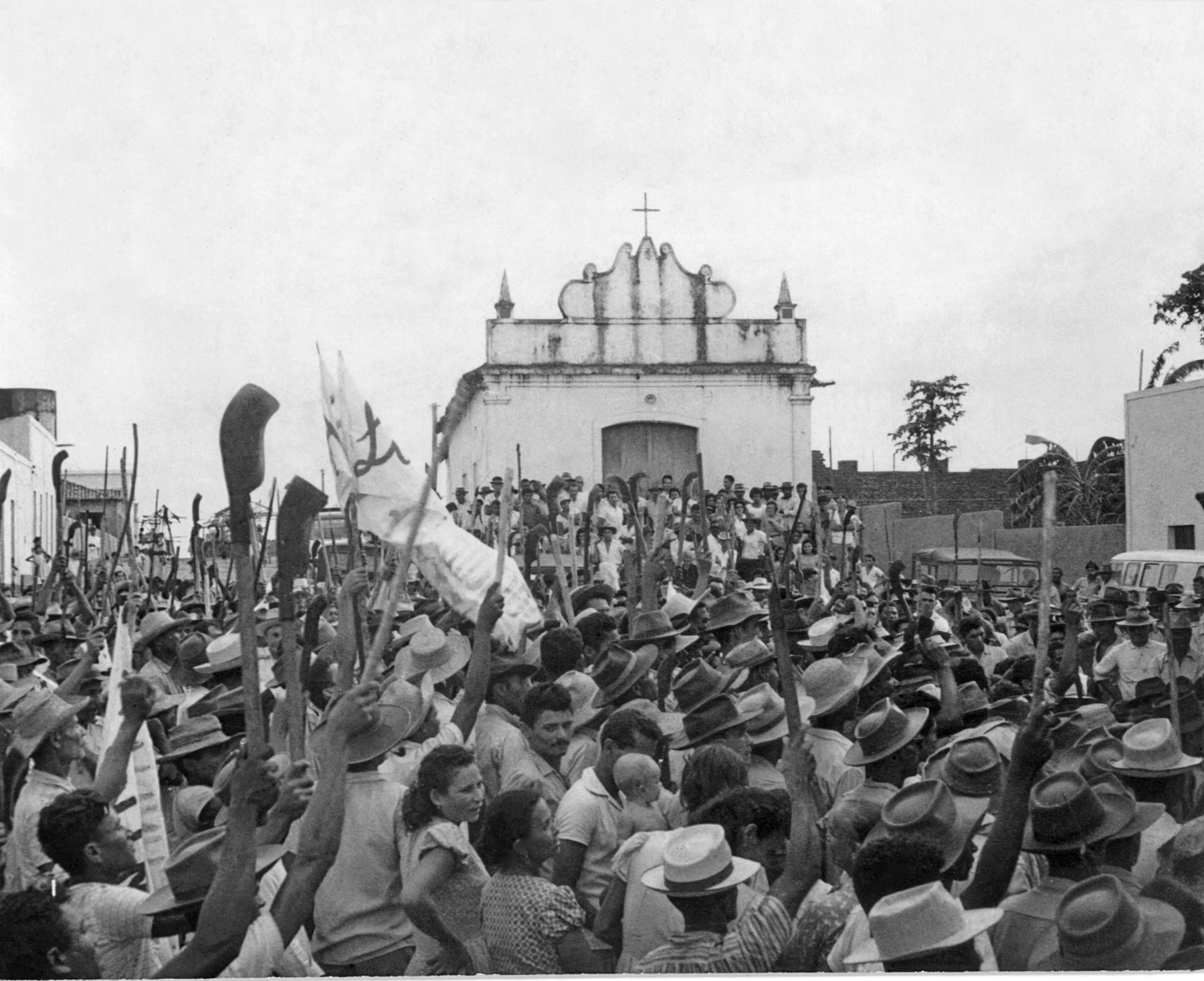 Fotografia em preto e branco. Homens e mulheres reunidos em um protesto em frente a uma pequena igreja. Estão erguendo ferramentas de trabalho no campo.