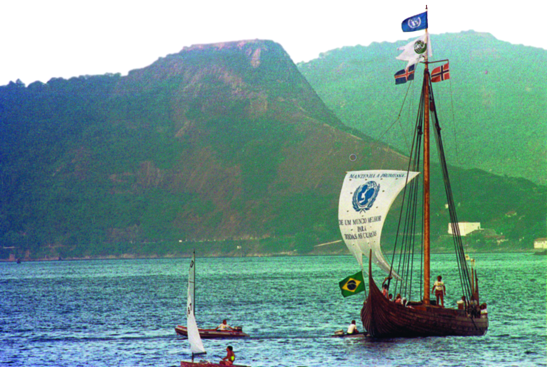 Fotografia. Sobre águas calmas de cor verde escura, à direita, uma embarcação à vela, feita de madeira, com um longo mastro com quatro bandeiras hasteadas: no alto, a bandeira da ONU; abaixo, uma bandeira branca com o logotipo internacional da Conferência das Nações Unidas sobre o Meio Ambiente de 1992 (a Rio -92); à esquerda, a da Islândia e à direita, a da Noruega. Amarrados ao topo do mastro, os cabos formam um triângulo no centro da embarcação. Há um estandarte branco amarrado na proa, com o emblema da UNICEF em azul e o texto Mantenha a promessa de um mundo melhor para todas as crianças. Acima do costado da embarcação, todo feito de madeira, a bandeira do Brasil. Um tripulante da embarcação está no centro do convés em pé. Outro acena com um braço para ao alto. À direita, há dois botes infláveis movidos a motor, cada um deles pilotado por um homem. Mais à frente, há um pequeno catamarã, visto de lado. Ao fundo, montanhas cobertas de vegetação verde e, no alto, o céu azul claro.