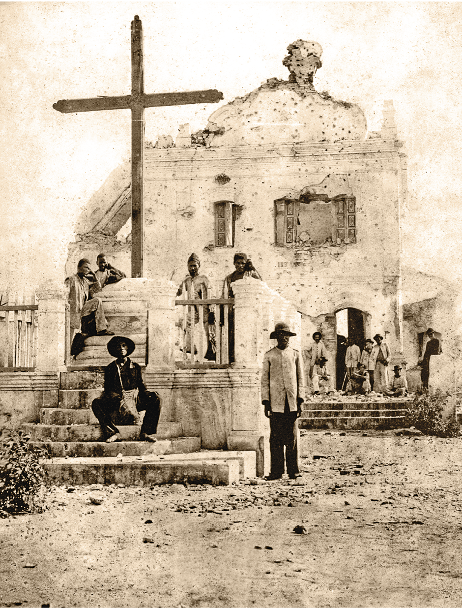 Fotografia em sépia. Pessoas em frente a uma igreja. A construção tem partes destruídas e muitas marcas de tiro. À frente, pessoas em pé e sentadas ao redor de uma grande cruz de madeira.