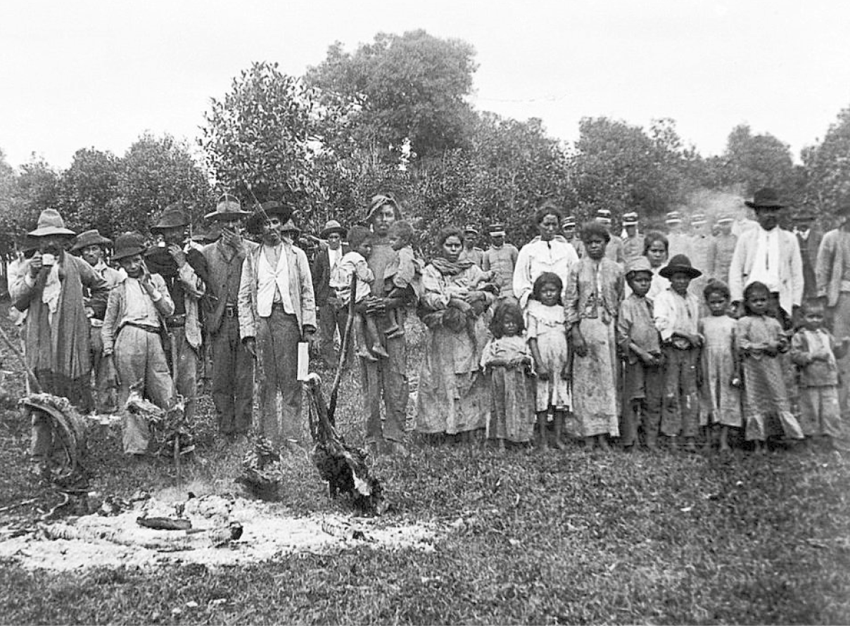 Fotografia em preto e branco. Famílias reunidas no campo. Há muitos homens, mulheres e crianças vestindo roupas simples. Ao fundo, perto da vegetação, militares.