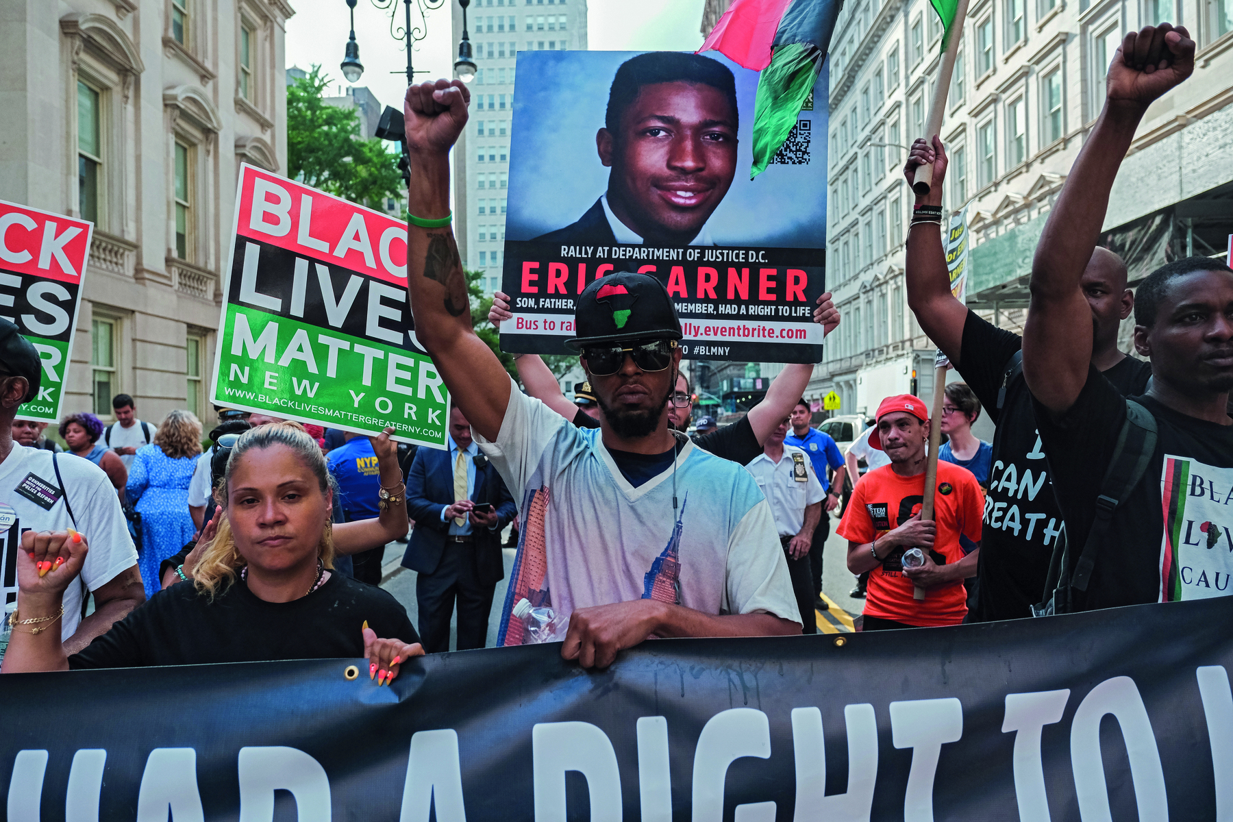 Fotografia. Grupo de pessoas em um protesto. Carregam cartazes com palavras de ordem. Em um dos cartazes a foto de Eric Garner, rapaz negro.