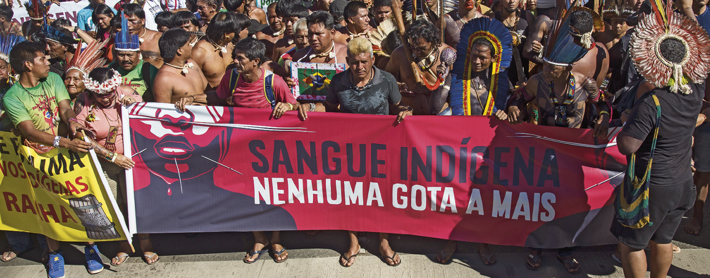 Fotografia. Indígenas reunidos em um protesto. Alguns usam cocares sobre a cabeça. À frente, um grande cartaz vermelho com os dizeres: SANGUE INDÍGENA, NENHUMA GOTA A MAIS.