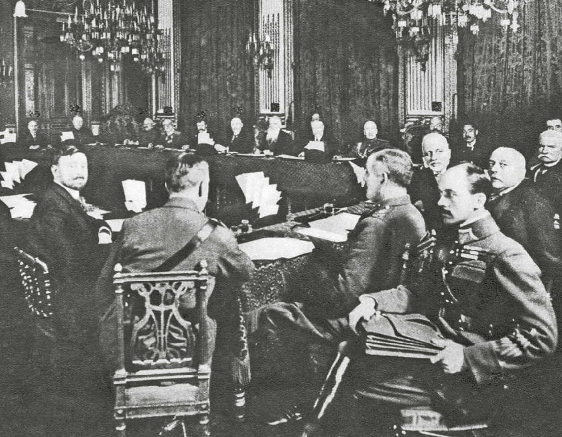 Fotografia em preto e branco. Homens reunidos em um salão amplo. Estão sentados em um grande círculo. Alguns usam roupas militares, outros, roupas sociais.