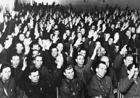 Fotografia em preto e branco. Grupo de militares sentados com os braços direitos erguidos.