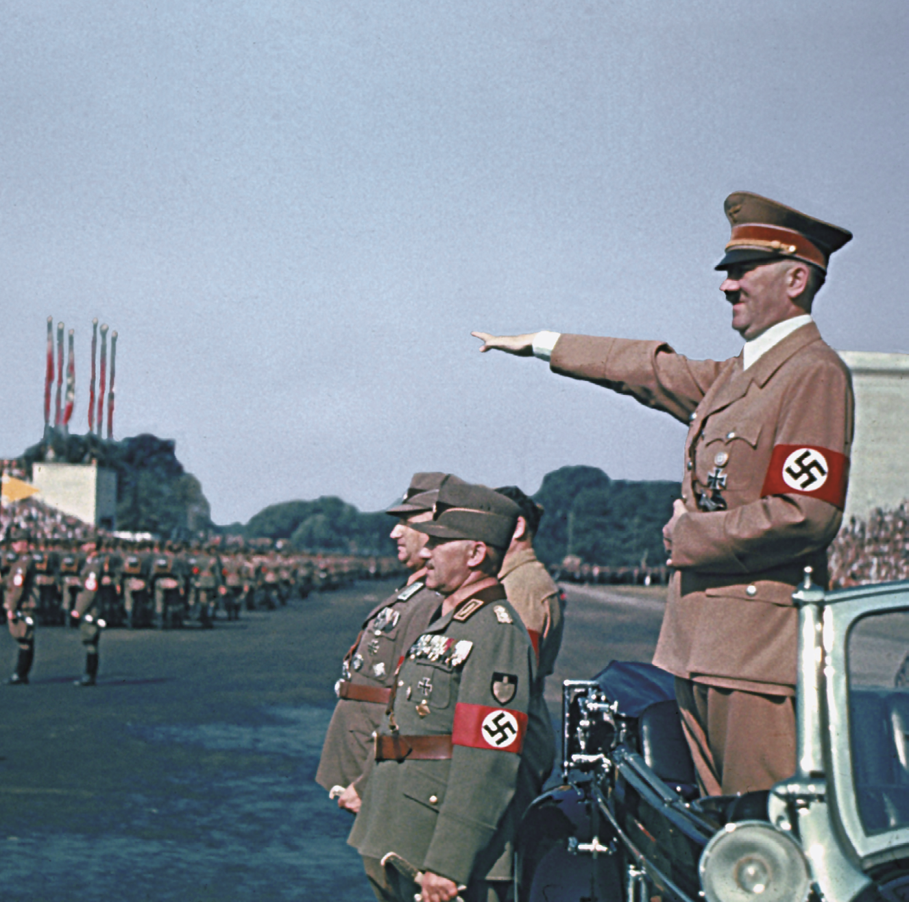 Fotografia. Adolf Hitler em pé, dentro de um carro com a capota aberta. Ele veste uniforme marrom claro. Em um dos braços uma braçadeira vermelha com o símbolo da suástica. Ele saúda os militares perfilados com o braço esticado e a palma da mão para baixo.