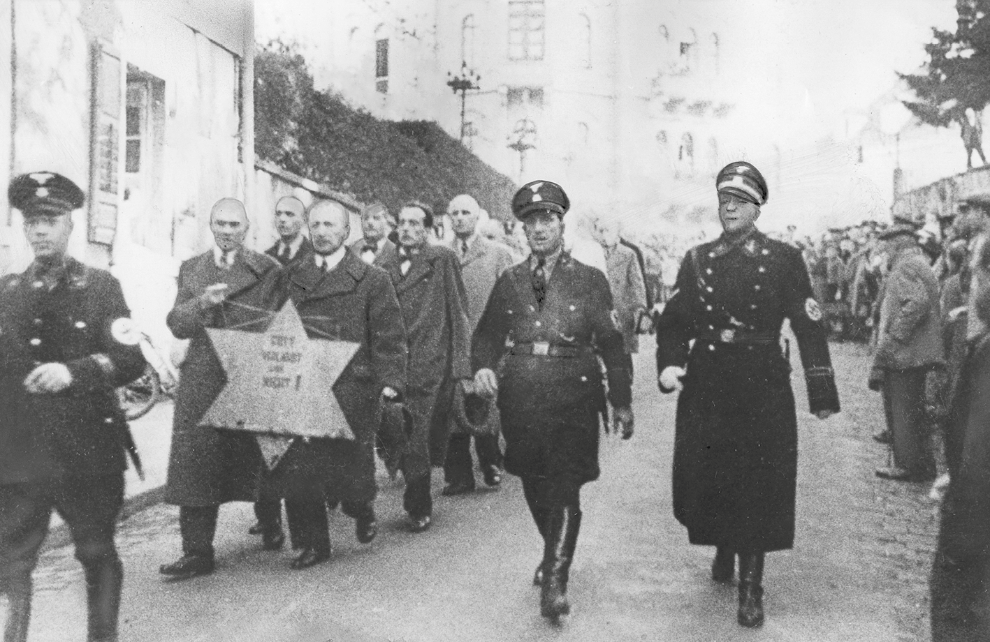 Fotografia em preto e branco. Judeus em duplas em uma longa fila. Caminham pela rua escoltados por militares. A dupla da frente carrega uma estrela de Davi, símbolo do judaísmo.
