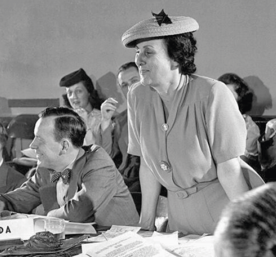 Fotografia em preto e branco. Mulher vestindo camisa, saia e chapéu. Está em pé com as mãos apoiadas sobre uma bancada.