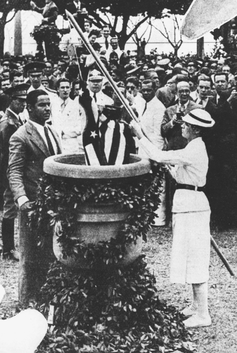 Fotografia em preto e branco. No centro da imagem, uma mulher de saia comprida e reta, casaco e chapéu claros, coloca uma bandeira dentro de um recipiente alto. Ao redor, diversos homens vestidos socialmente observam a cena.
