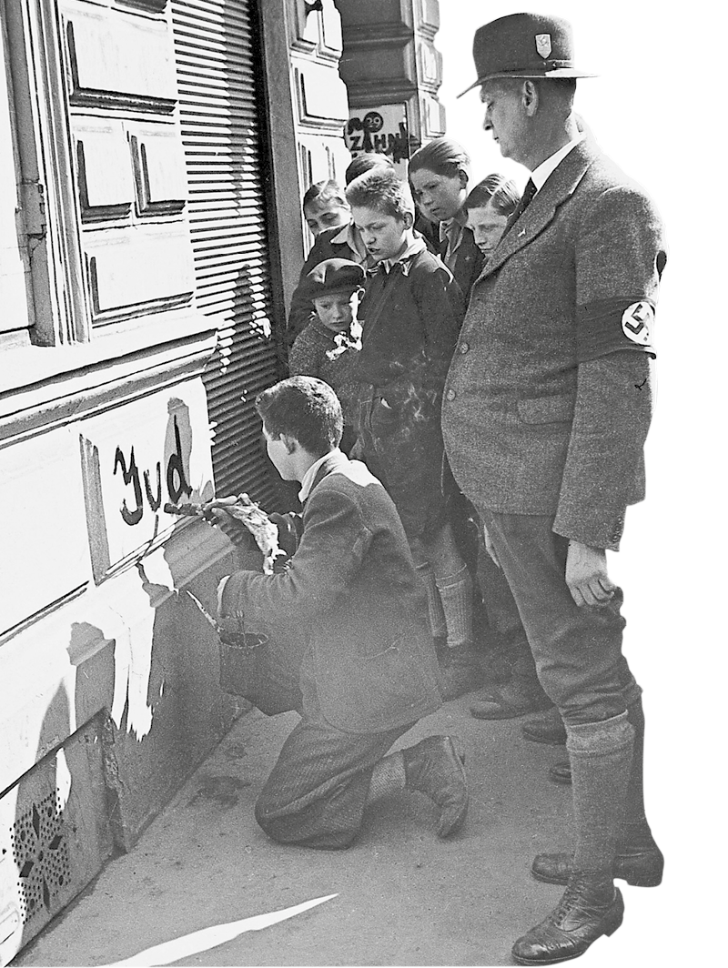 Fotografia em preto e branco. Rapaz de roupas sociais abaixado escrevendo em um muro. Ao redor dele outros jovens e um homem com uma braçadeira nazista.