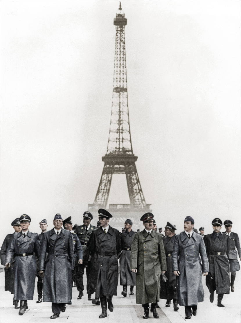 Fotografia colorizada. No primeiro plano, Adolf Hitler com um longo casaco verde e quepe escuro. Está caminhando ao lado de outros homens de casaco e quepe. Atrás, militares agrupados. Ao fundo a Torre Eiffel.