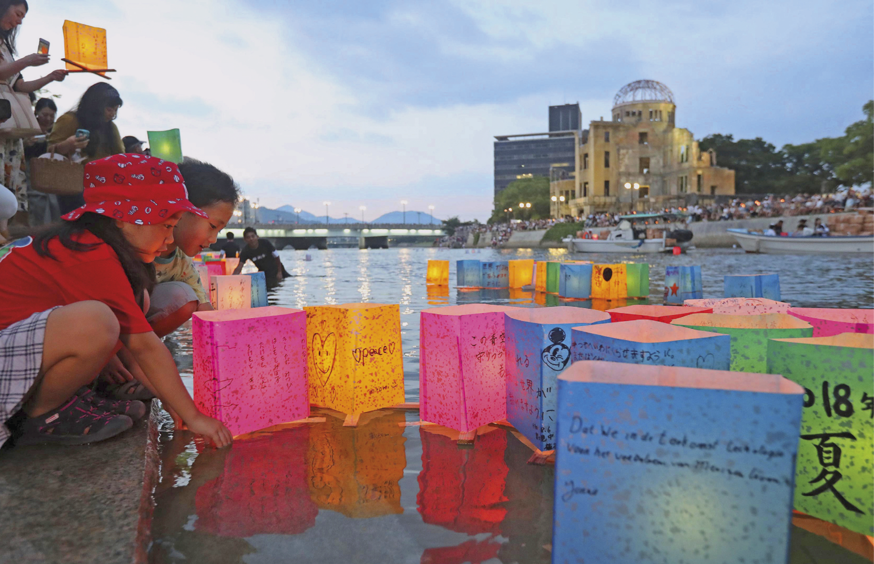 Fotografia. No primeiro plano, crianças e adultos colocando lanternas coloridas de papel em um grande rio. Diversas pessoas nas margens observam. Ao fundo duas construções, uma está em ruínas.