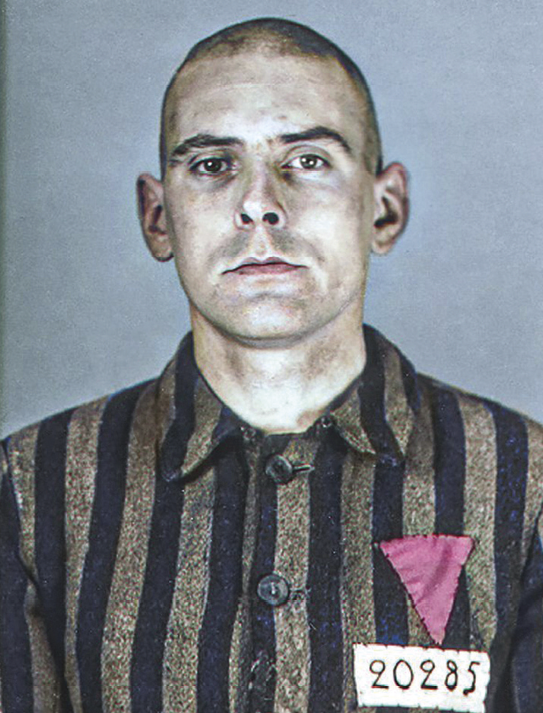 Fotografia. Homem de rosto comprido, cabelo raspado, orelhas grandes e queixo quadrado. Veste uma camisa com listras verticais. No canto da camisa um número e um triângulo rosa de ponta cabeça.