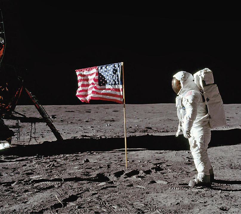 Fotografia. Astronauta pisando no solo lunar. Ele veste macacão, botas, luvas e capacete. À sua frente, a bandeira dos Estados Unidos em um pequeno mastro fincado no chão. O solo é escuro e irregular.
