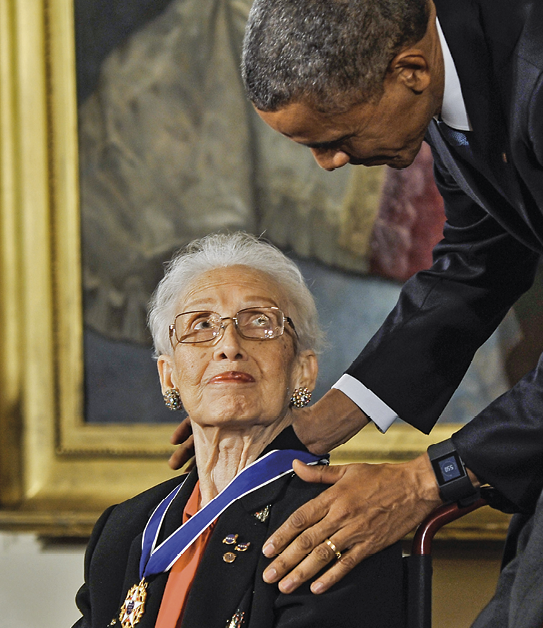 Fotografia. Barack Obama, homem de cabelo grisalho, está abaixado com as mãos nos ombros de Katherine Johnson, senhora de cabelo branco, olhos pequenos e óculos. Ela está com a cabeça inclinada para trás, olhando para Obama. Ela tem uma medalha pendurada no pescoço.