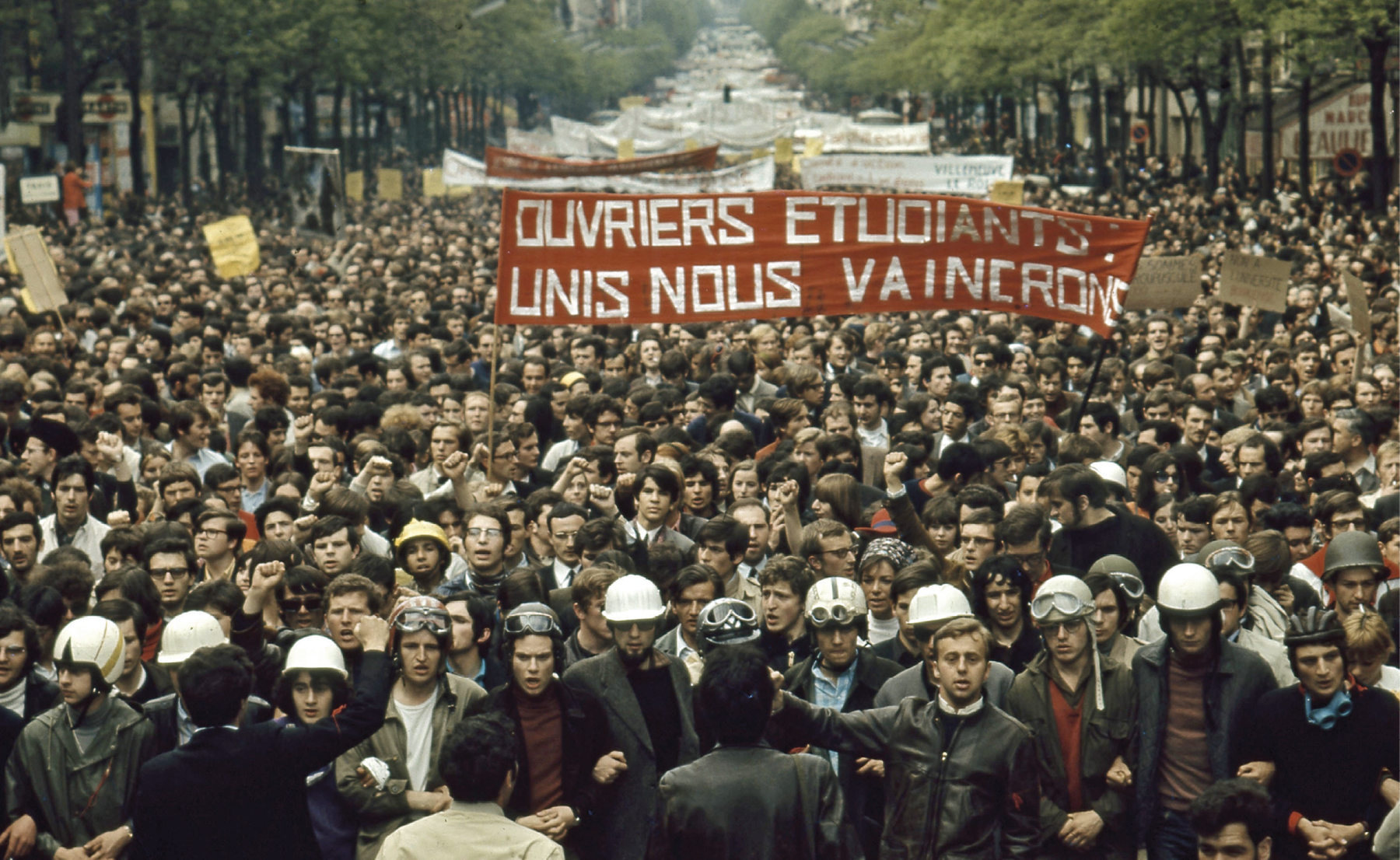 Fotografia. Milhares de pessoas em uma manifestação ocupam uma longa avenida. Carregam faixas e cartazes. Na primeira fila estão lado a lado, de braços dados. No centro, uma faixa vermelha com os dizeres em francês.