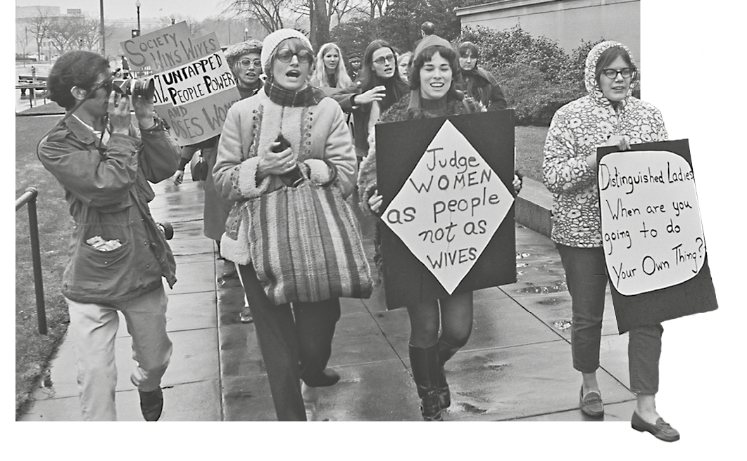 Fotografia em preto e branco. Mulheres em uma manifestação. Carregam cartazes feitos à mão. À esquerda, um homem fotografa a passeata.