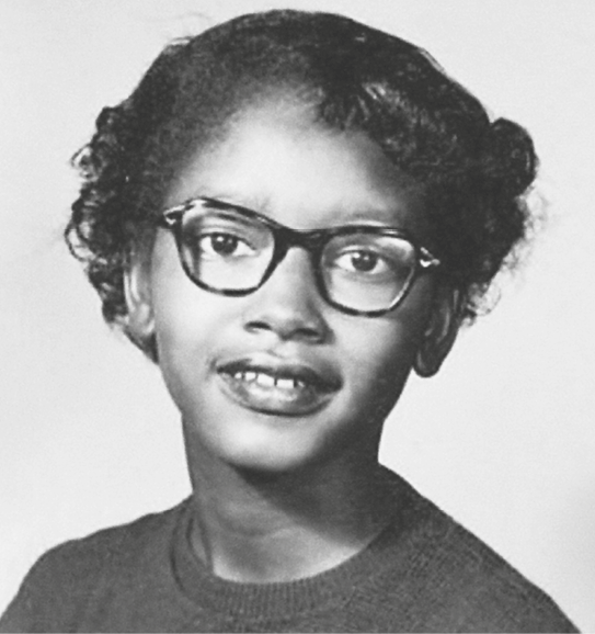 Fotografia em preto e branco. Menina negra, de cabelos curtos. Usa óculos de armação quadrada.