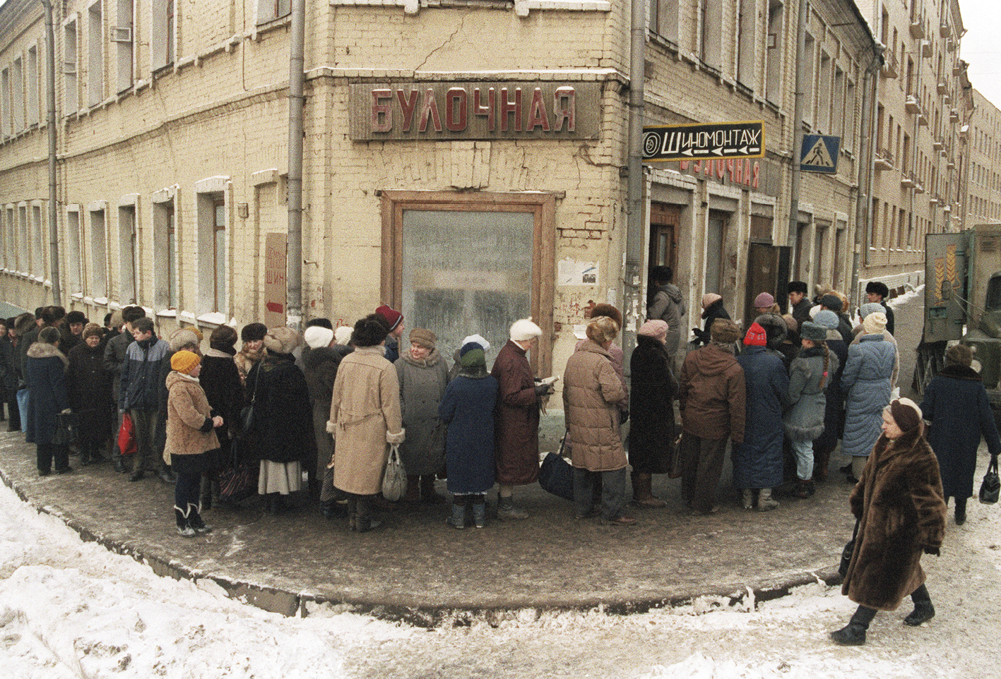 Fotografia. Pessoas em uma longa fila em um estabelecimento de esquina. Todos estão de casaco e touca ou chapéu de frio. Na rua há neve.