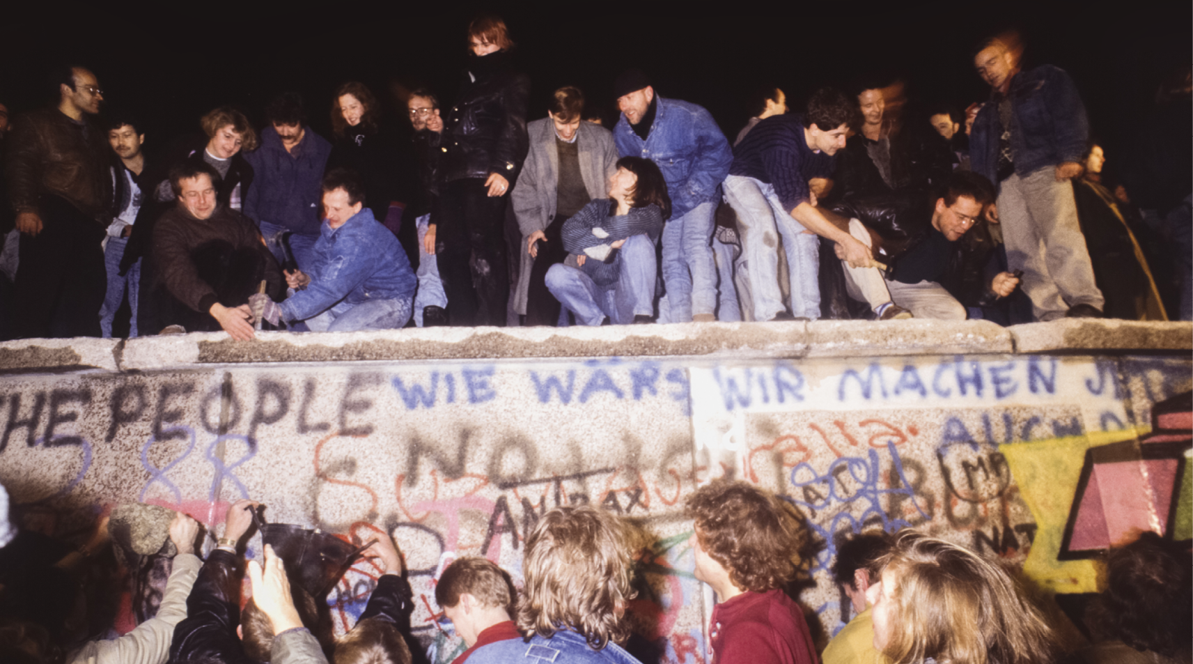 Fotografia. Pessoas no topo do muro de Berlim. À frente do muro, outras pessoas observam. Ele está pichado com símbolos e frases em inglês e alemão.