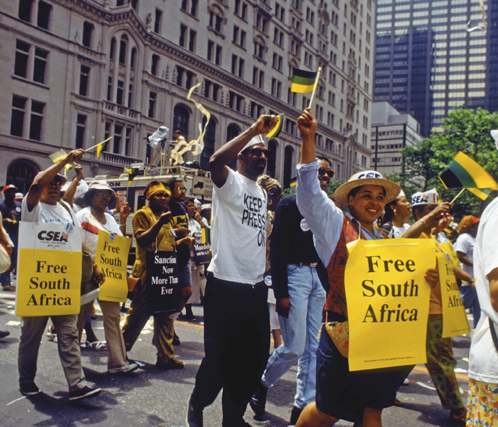 Fotografia. No primeiro plano, pessoas em uma manifestação na rua. Carregam cartazes amarelos com palavras de ordem em inglês pendurados ao corpo.