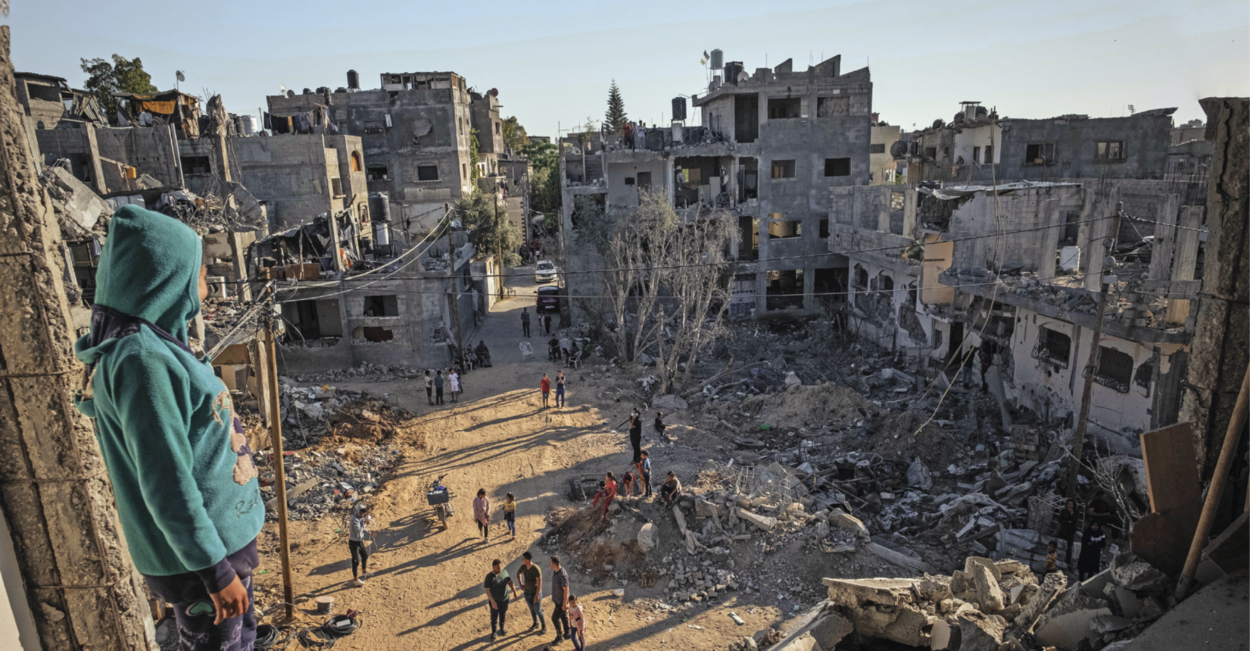 Fotografia. Em primeiro plano, mulher no alto, observa as casas e prédios em ruínas. No centro da imagem, algumas pessoas estão entre os escombros, observando o que restou do local.