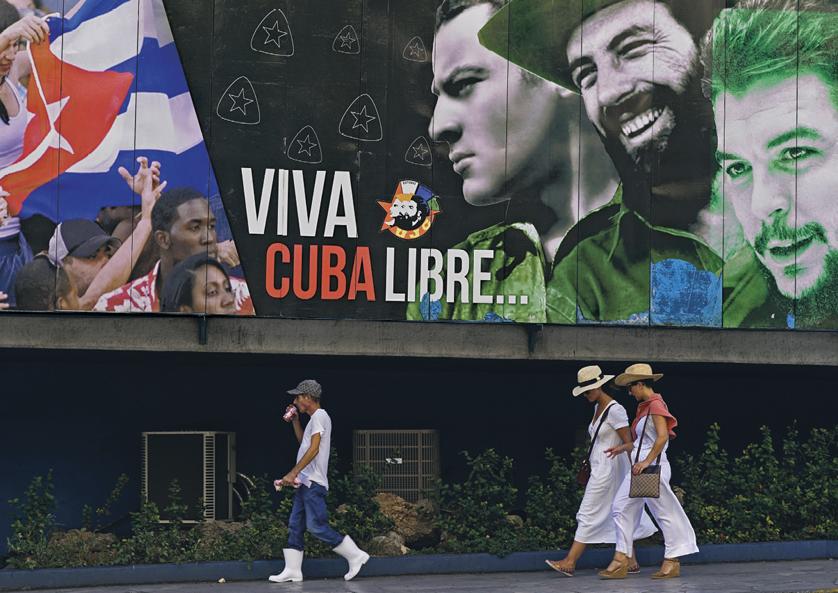 Fotografia. No primeiro plano, três pessoas passando em uma calçada. No segundo plano, um mural representando, à direita, três homens sorridentes e, à esquerda, pessoas aglomeradas carregando uma bandeira de Cuba. No centro do mural, uma frase cuja tradução é Viva Cuba livre...