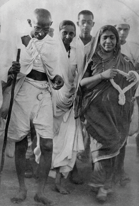 Fotografia em preto e Branco. Mahatma Gandhi, senhor de óculos, mago, careca, com o corpo envolto em um tecido claro. Está descalço. Se apoia em um cajado. Atrás dele, homens e mulheres.