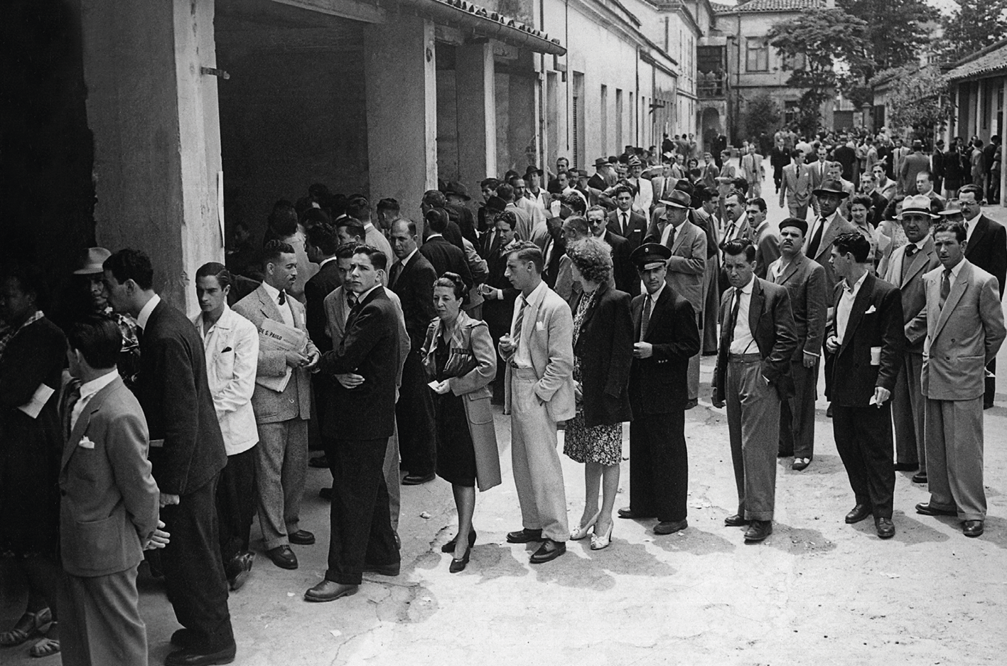 Fotografia em preto e branco. Homens e mulheres trajados socialmente dispostos em filas na porta de um edifício.