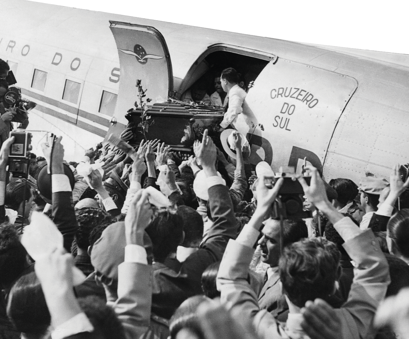 Fotografia em preto e branco. Homens apoiando um caixão na porta de um avião. Ao redor uma multidão. Algumas pessoas fazem registro de imagens, outras acenam.