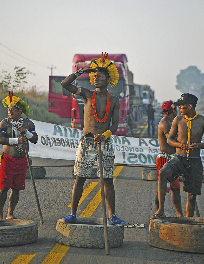 Fotografia. No primeiro plano, vistos de frente, indígenas de bermuda, alguns vestem cocar outros, boné. Estão se manifestando em uma estrada, em cima de pneus.