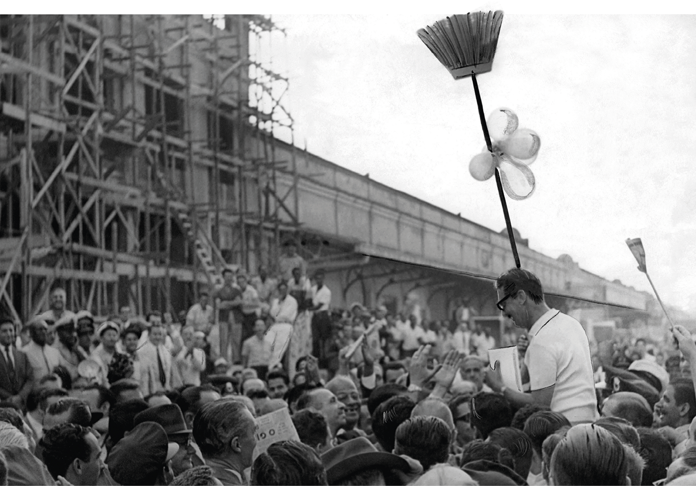 Fotografia em preto e branco. Jânio Quadros sendo carregado, no meio da multidão. Ao fundo, duas vassouras levantadas para o alto.