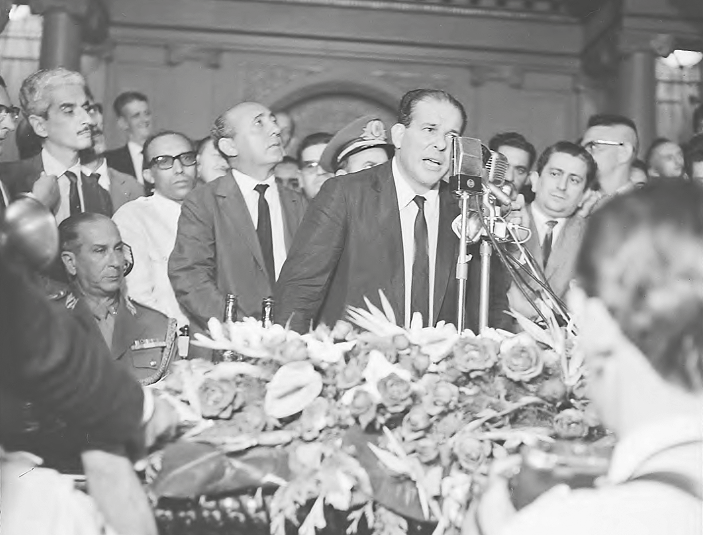 Fotografia em preto e branco. João Goulart, de terno e gravata, cabelos penteados para trás. Está em pé, falando em um microfone. Ao seu redor, diversos homens acompanham o ato.