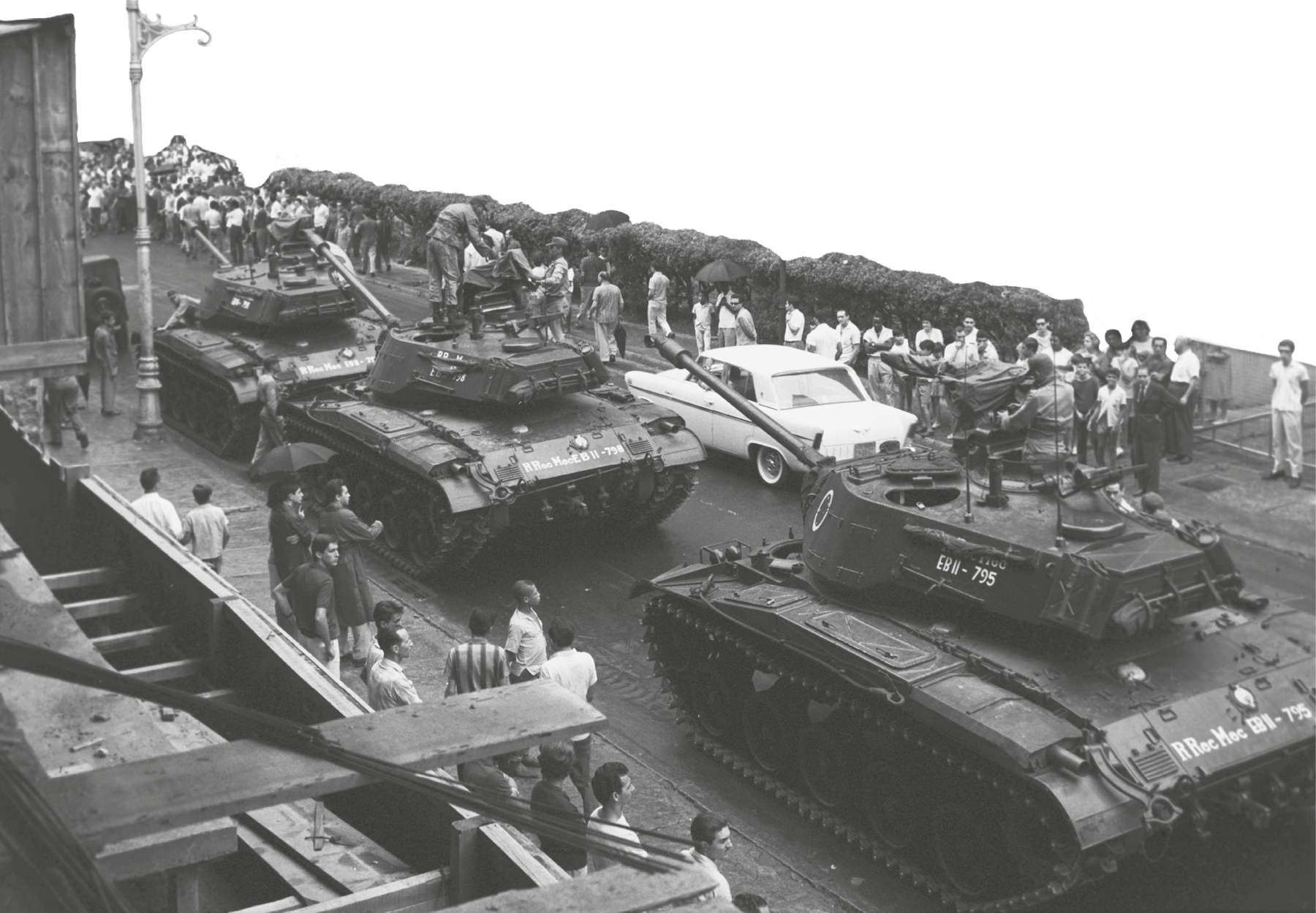 Fotografia em preto e branco. Tanques de guerra passando em uma avenida em meio a pessoas nas calçadas acompanhando o ato.
